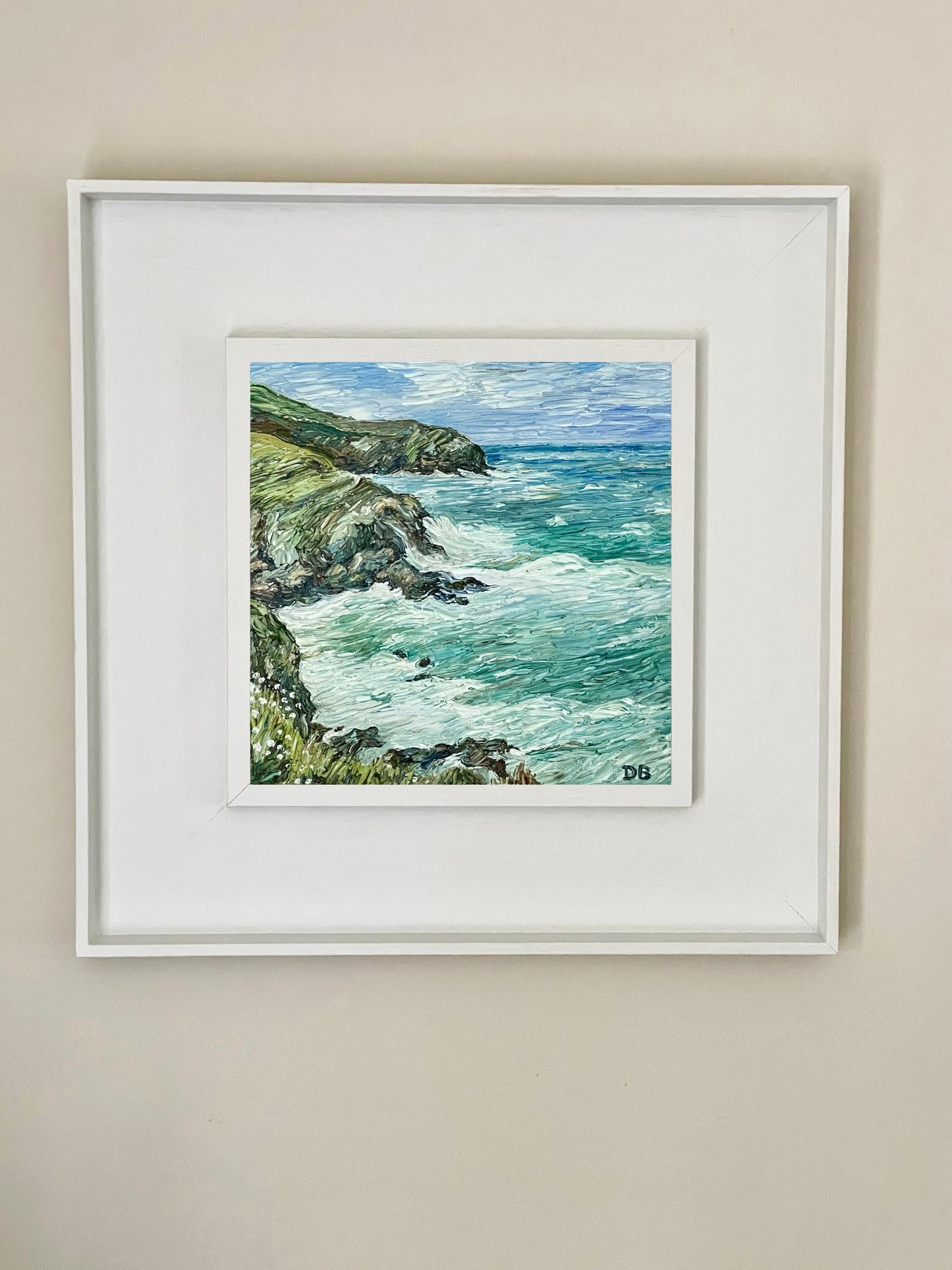 Framed artwork of a coastal scene with cliffs and ocean on a beige wall.